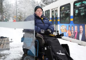 Person in power wheelchair, bundled up for winter weather, snow on the street, at bus shelter next to transit bus.