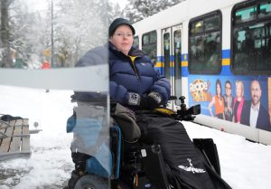 Person in power wheelchair, bundled up for winter weather, snow on the street, at bus shelter next to transit bus.