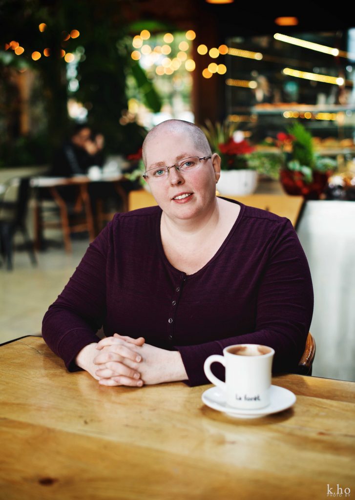 Photograph of Heather McCain, a bald, freckled non-binary person with glases. They are sitting at a table, hands crossed, beside them is a hot chocolate, in the background is a La Forêt, a cafe.