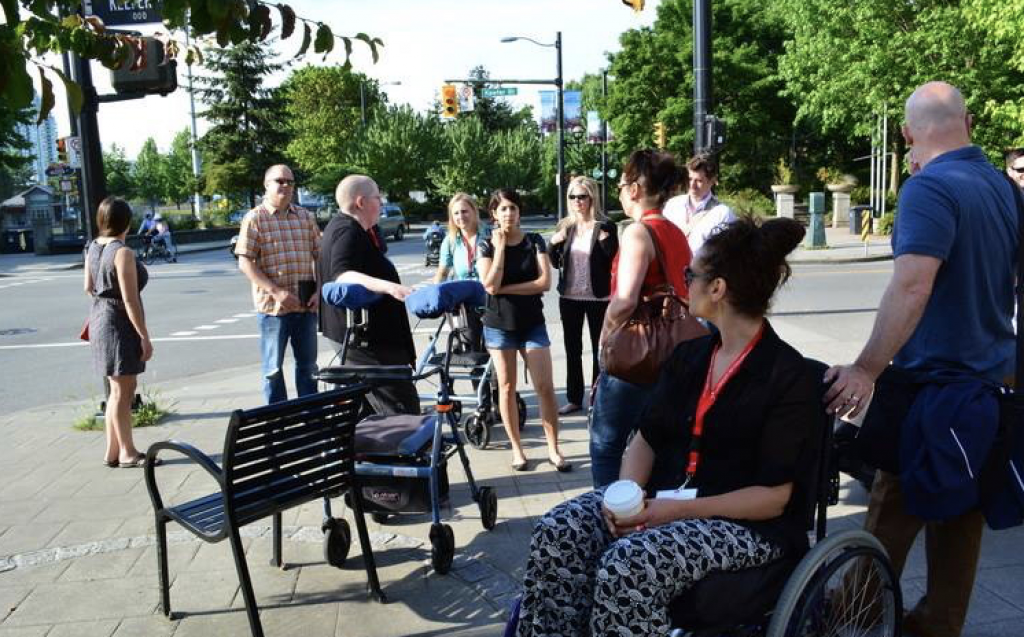 A group of people on an accessibility tour in downtown Vancouver. 
The group is comprised of people of various ages, genders, sexualities, disabilities, and races. 